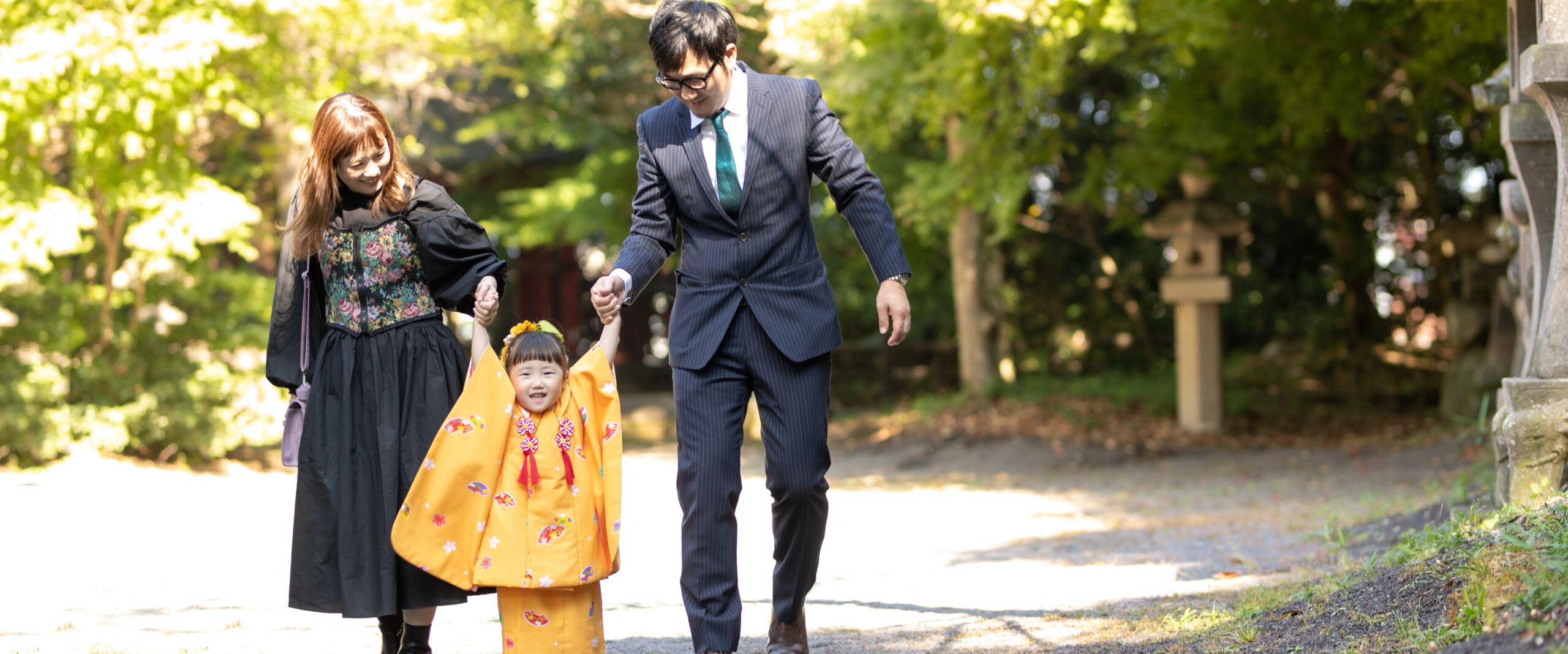 中津の薦神社の豊かな緑に囲まれた参道で、黄色の着物を着た女の子がパパとママに両手を引かれて笑顔で歩く七五三家族写真