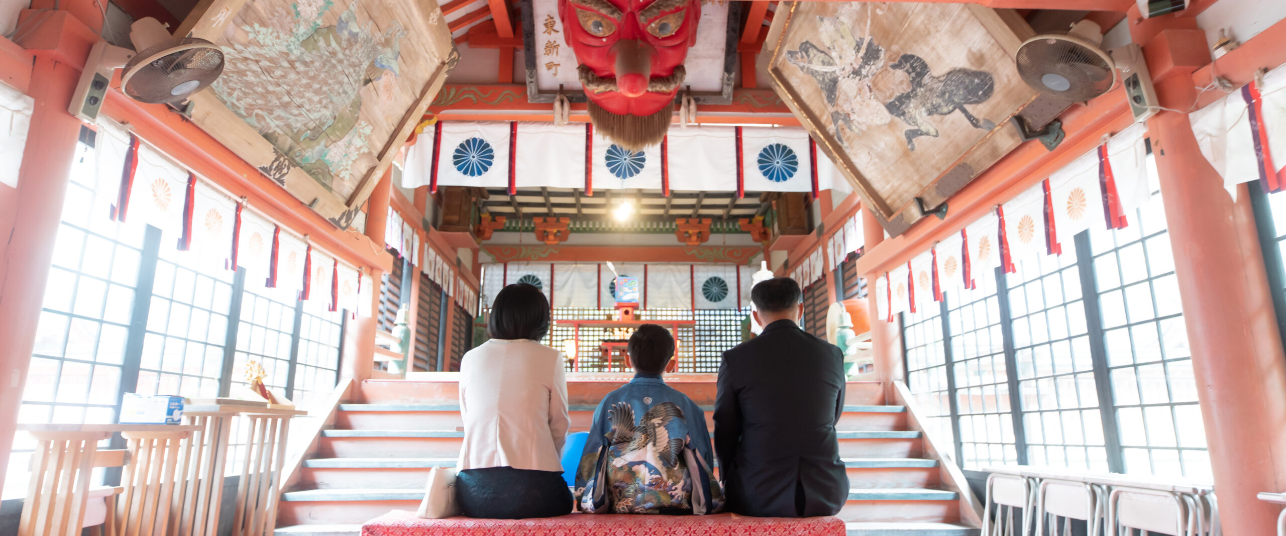 大分の柞原八幡宮の拝殿に掲げられた大きな天狗の面の下で、お参りに訪れたご家族が静かに並んで座る七五三の後ろ姿