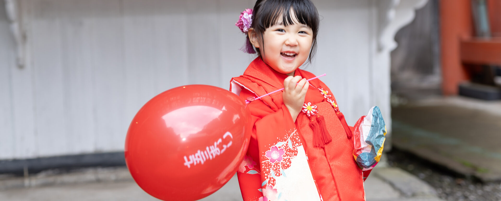 **代替テキスト:**
大分市の柞原八幡宮で、赤い着物を着た3歳の女の子が風船を持って笑顔を見せる七五三のお参り写真。