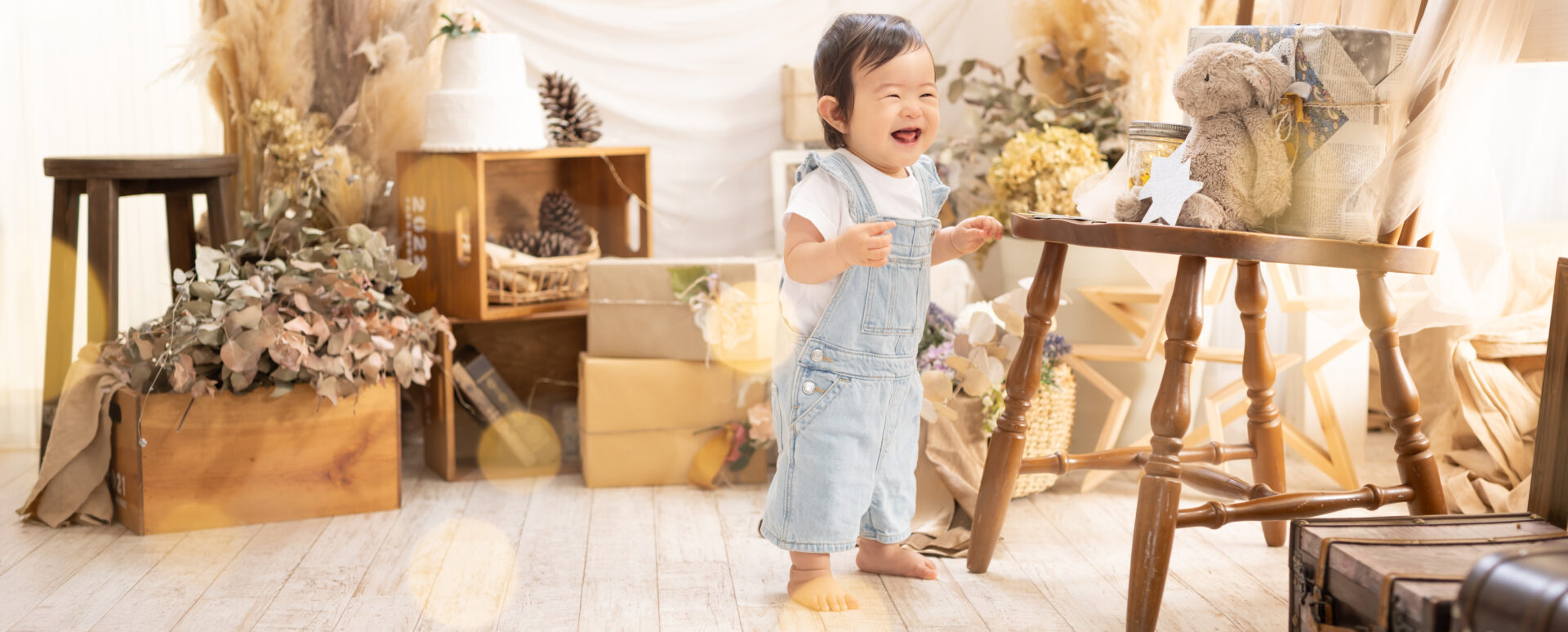 大分市のPhotostudio PASSIONで1歳記念撮影。白いTシャツとブルーのオーバーオールを着て、スタジオでつかまり立ちをしながら笑顔を見せる赤ちゃん。