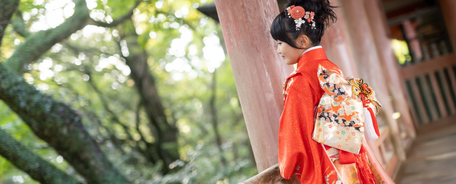 大分市で七五三のお参り撮影。赤い着物に華やかな帯と髪飾りを合わせた7歳女の子が、神社の社殿でたたずむ自然な姿を残したPhotostudio PASSIONの七五三ロケーション写真。