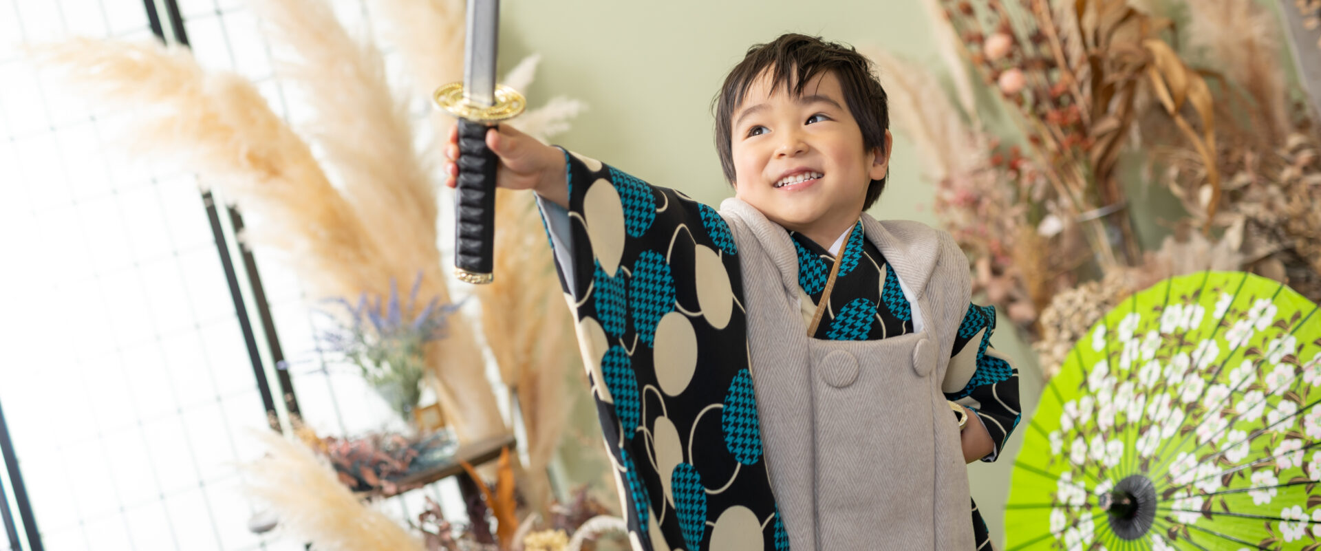 七五三の3歳男の子が、スタジオパッションで青とグレーの被布を着用し、刀を構えて笑顔でポーズをとる様子