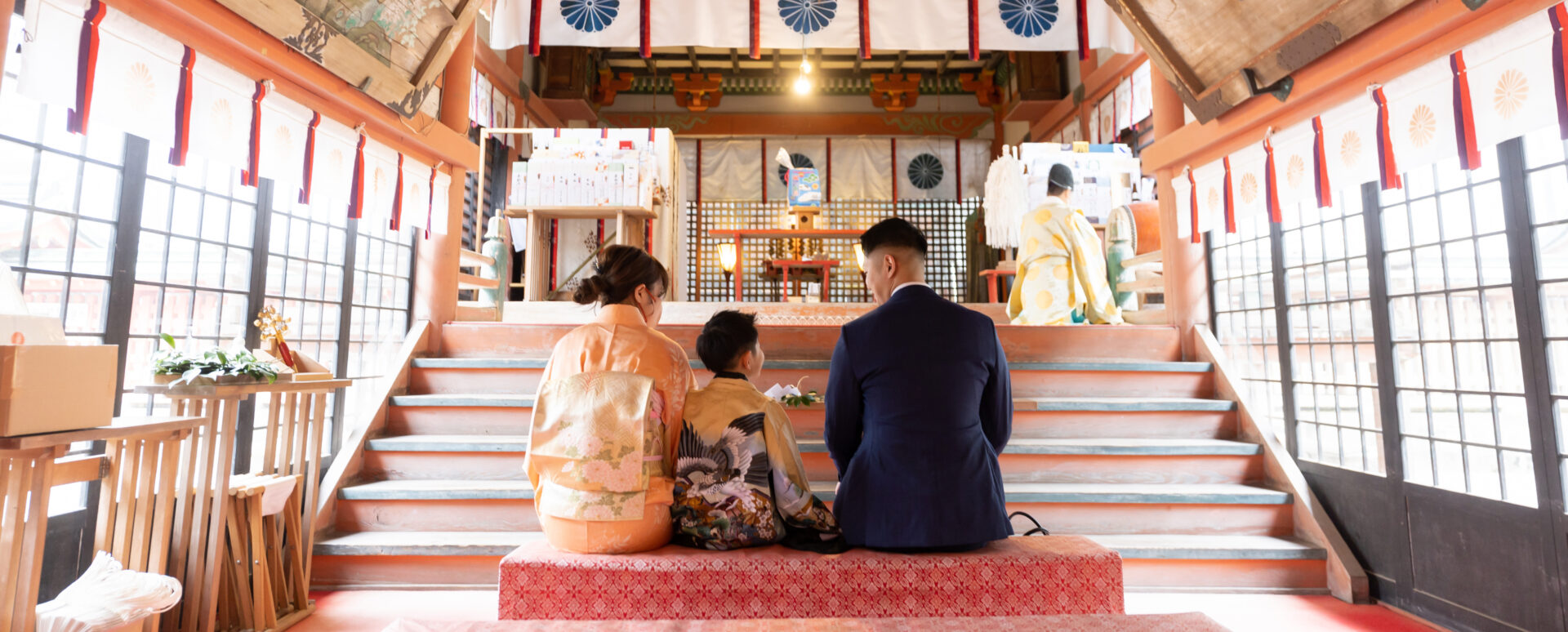 大分市の柞原八幡宮拝殿内で、菊の御紋や朱色の柱を背景に、鶴が描かれた羽織袴を着た男の子と和装の母親、スーツ姿の父親が並んで座る七五三ご祈祷の家族後ろ姿写真