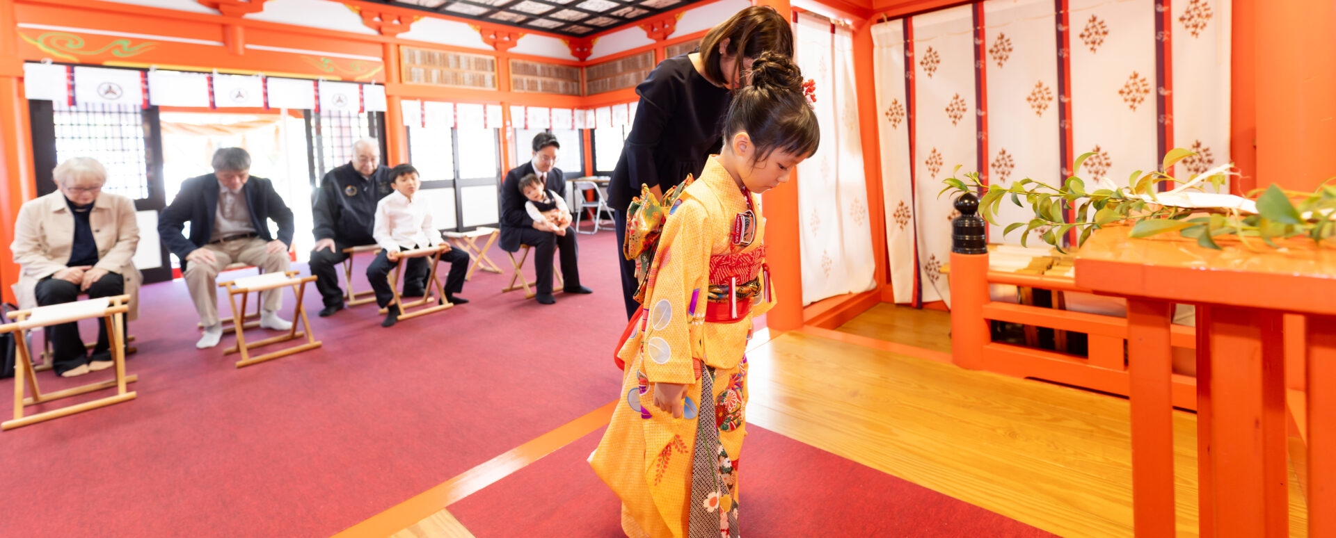 大分市の若宮八幡宮拝殿内で、鮮やかな朱塗りの柱を背景に、黄色の花柄着物を着た女の子が神前で深く一礼をし、後ろでご家族・ご親族が温かく見守る七五三ご祈祷写真