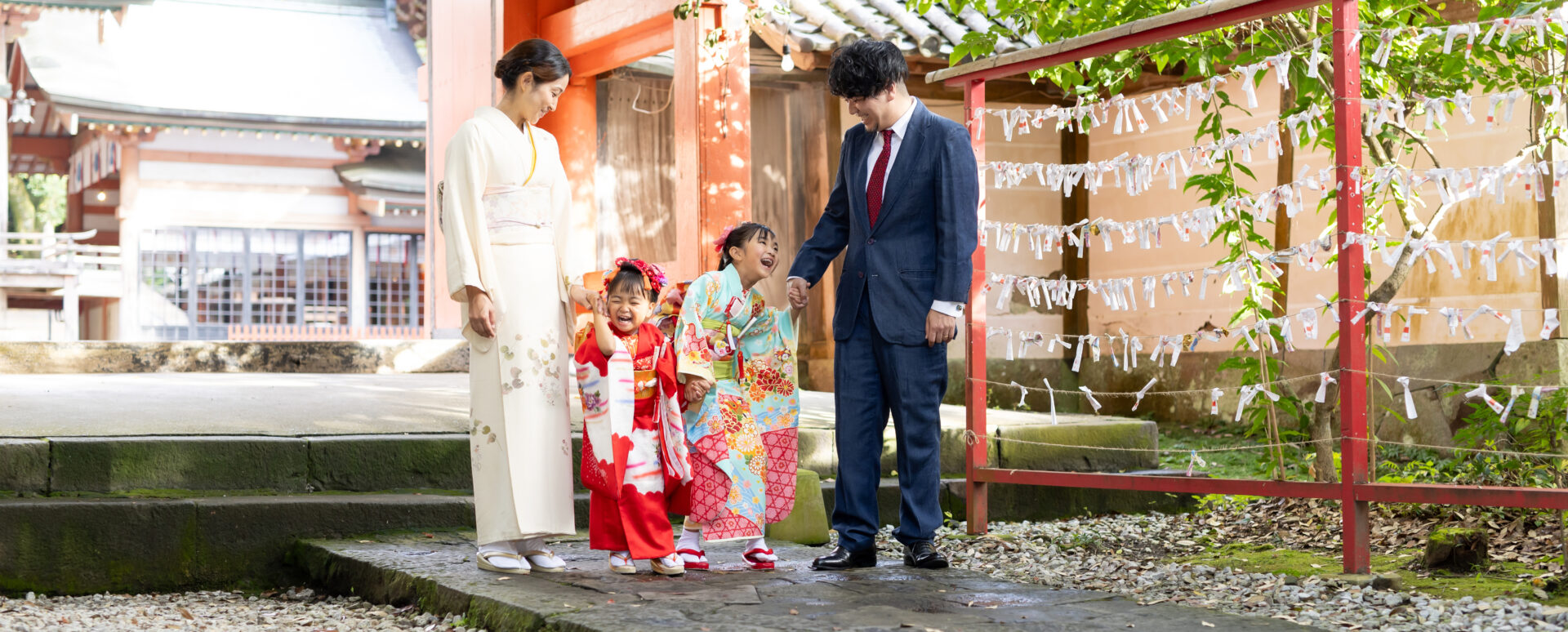 大分市の柞原八幡宮境内でおみくじ結び所を背景に、赤被布の3歳妹と水色着物の7歳姉の姉妹が顔を見合わせて大笑いし、和装の母親とスーツ姿の父親が優しく見守る七五三の家族写真