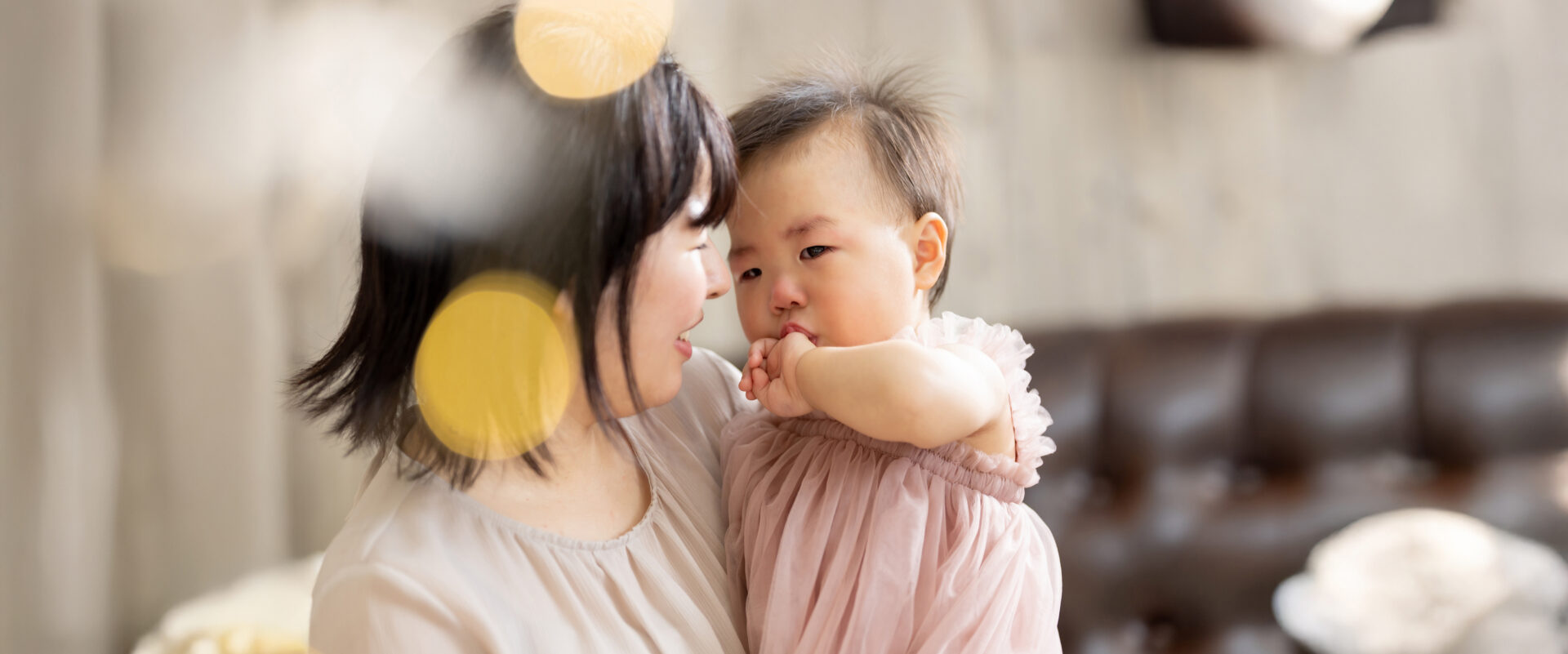 大分のスタジオ記念撮影で、優しい光のボケに包まれるなか、くすみピンクのドレスを着てママに抱っこされながら、甘えるように泣きそうな顔で見つめ合う親子写真