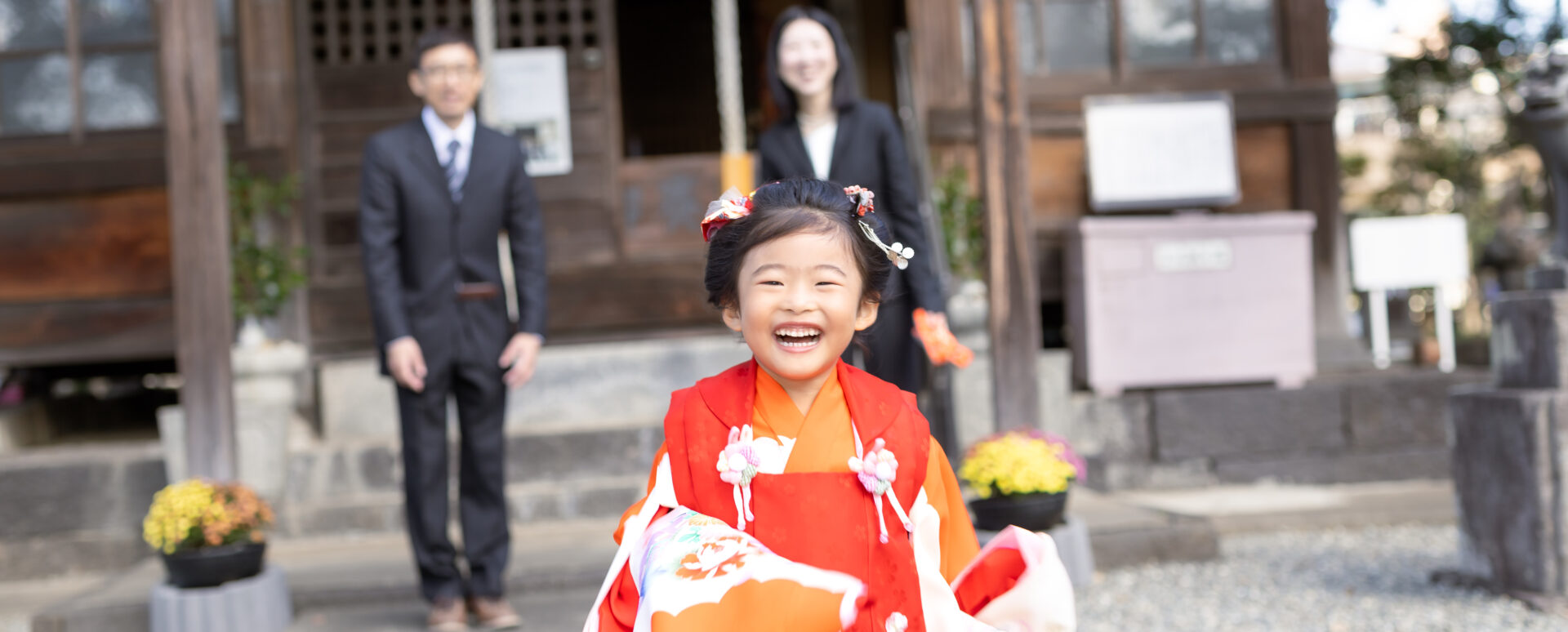 大分社の木造社殿のある神社境内で、鮮やかなオレンジの着物と赤い被布を着た女の子が満面の笑顔で手前を駆け寄り、後ろでスーツ姿のご両親が見守る七五三ロケーション写真