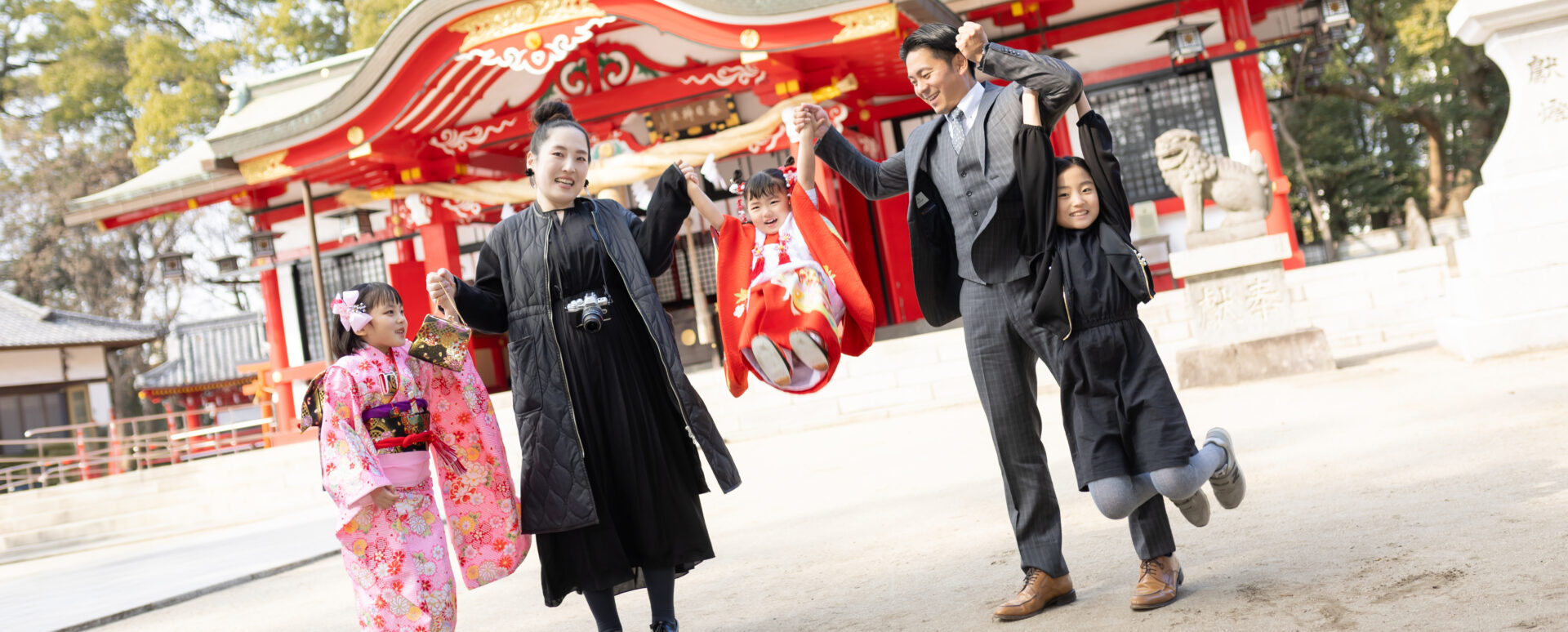 大分市の春日神社社殿前で、赤い被布を着た3歳女の子をご両親が持ち上げ、ピンクの着物を着た姉妹やスーツ姿の父親、カメラを掛けた母親の家族5人が笑顔で手を繋ぐ七五三写真