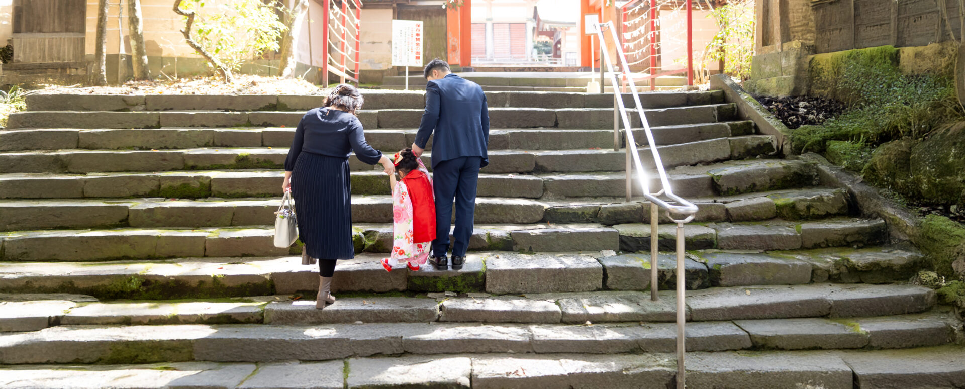 大分市の柞原八幡宮の苔むした石段参道で、白地にピンク花柄の着物と赤被布を着た3歳女の子がシックな服装のご両親と手を繋ぎ階段を登る七五三後ろ姿写真