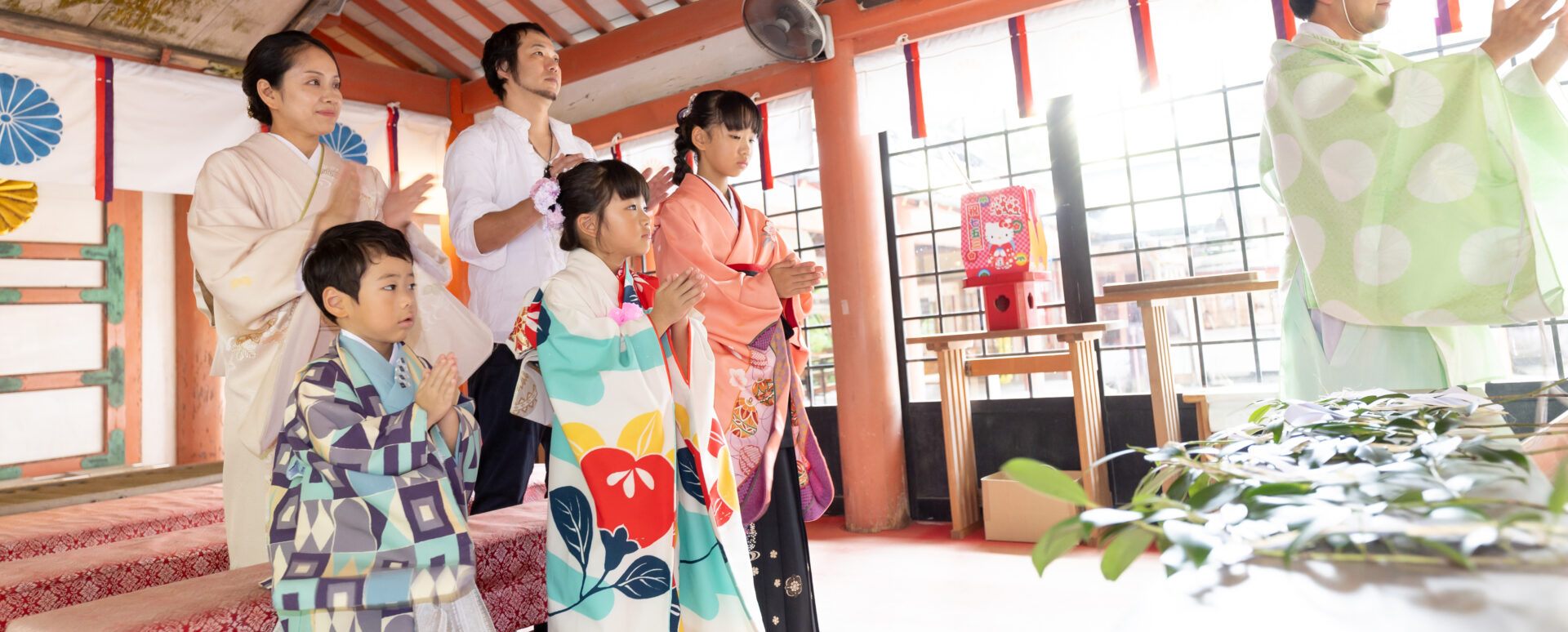 大分市の柞原八幡宮拝殿内で、レトロ柄の羽織袴を着た男の子とモダン椿柄の被布着物の女の子、ピンク着物のお姉ちゃんと和装の母親ら家族5人が神職様の前で静かに手を合わせる七五三ご祈祷写真