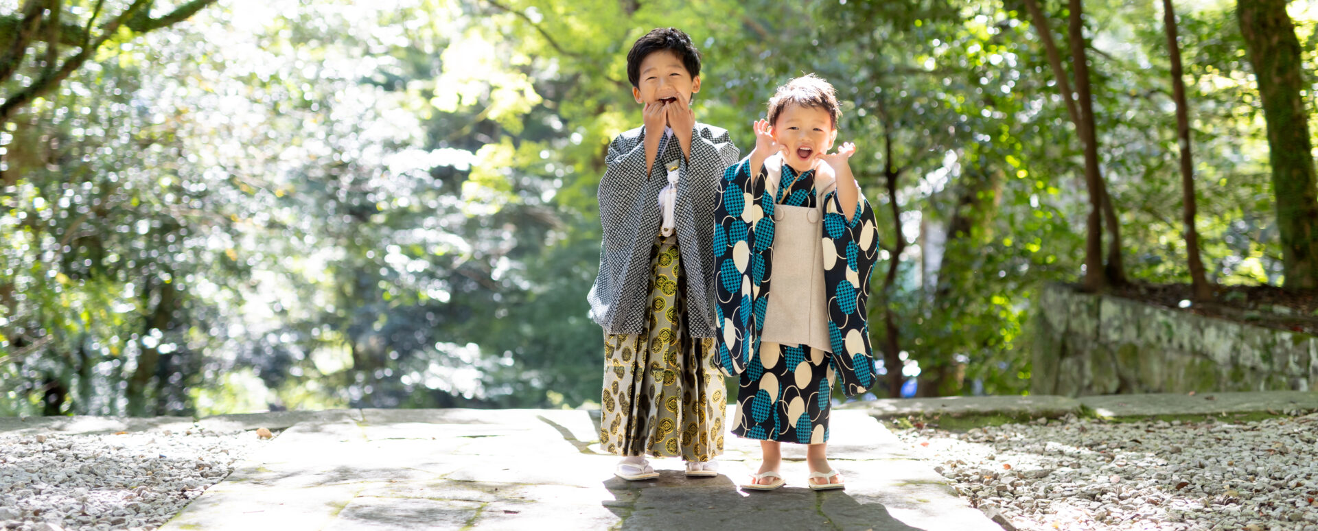 大分柞原八幡宮木漏れ日が差し込む神社の緑豊かな境内で、豪華な羽織袴を着た兄とモダン柄の被布着物を着た弟の兄弟が元気いっぱいにお茶目なポーズと笑顔を見せる七五三写真
