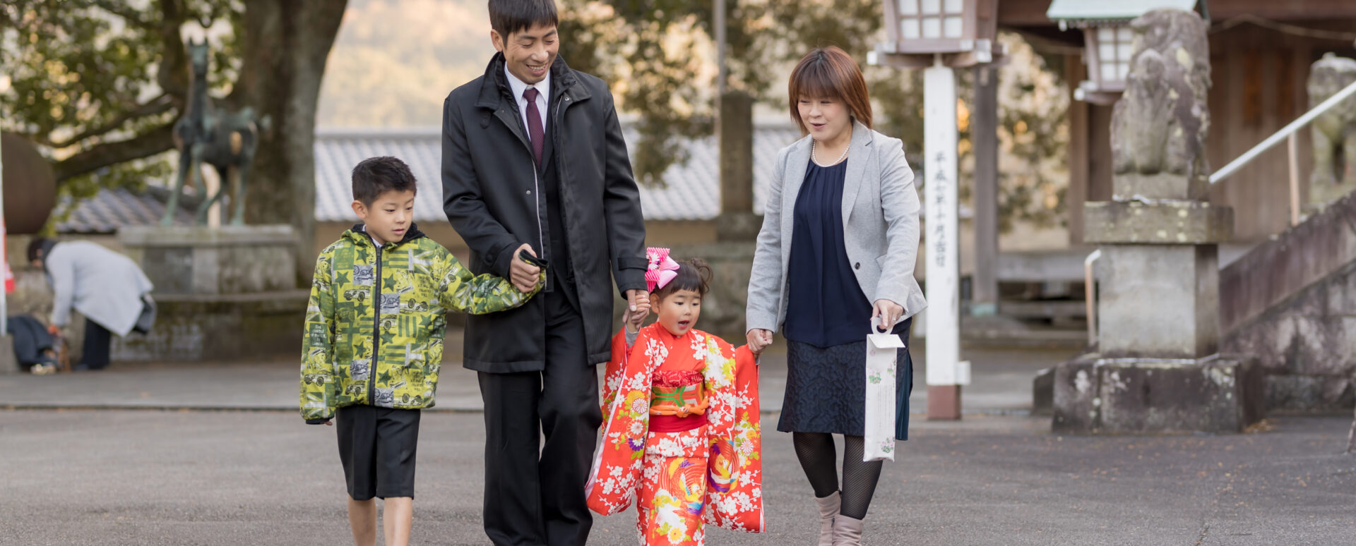 別府市の八幡竈門神社で七五三のお参りをする3歳の女の子。赤い着物姿でパパとママに手をつないでもらい、お兄ちゃんと一緒に歩く自然な家族写真。