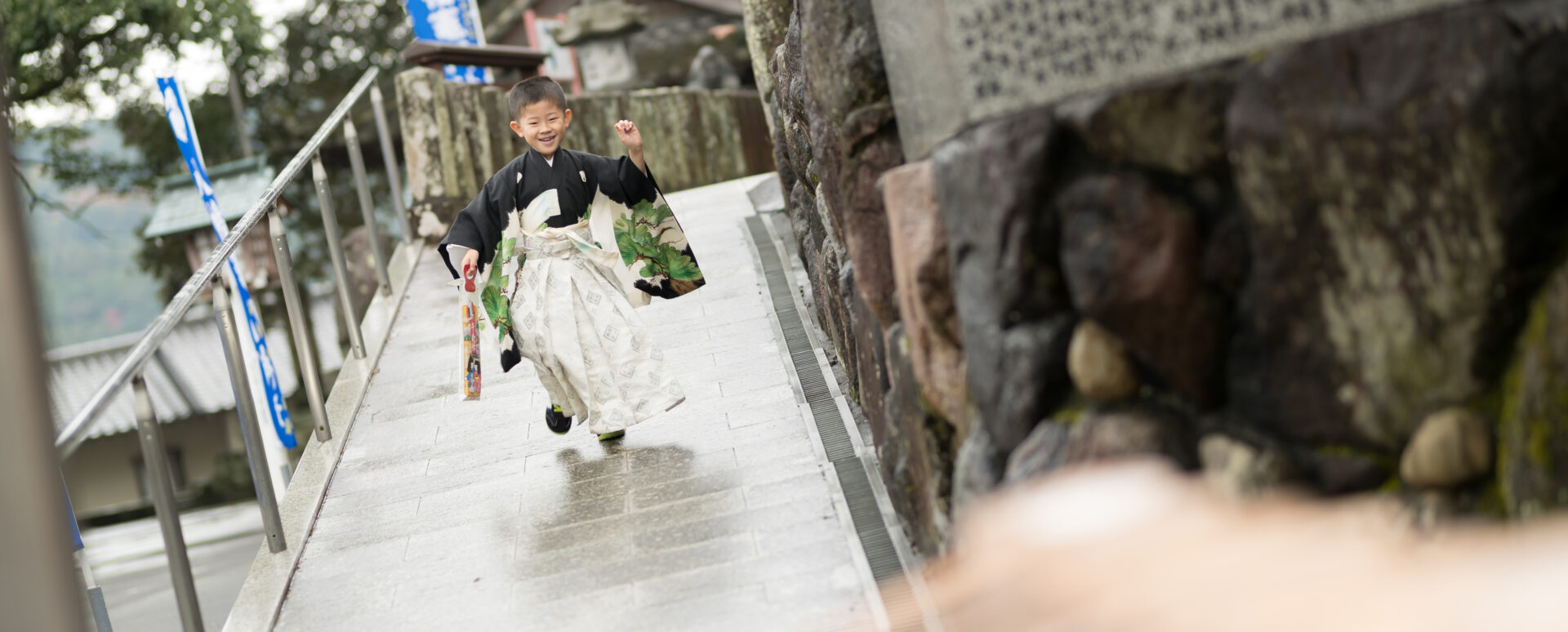 別府市の八幡竈門神社で七五三を迎えた5歳の男の子。黒と白の羽織袴姿で、千歳飴を手に笑顔で参道を走る自然な七五三写真。