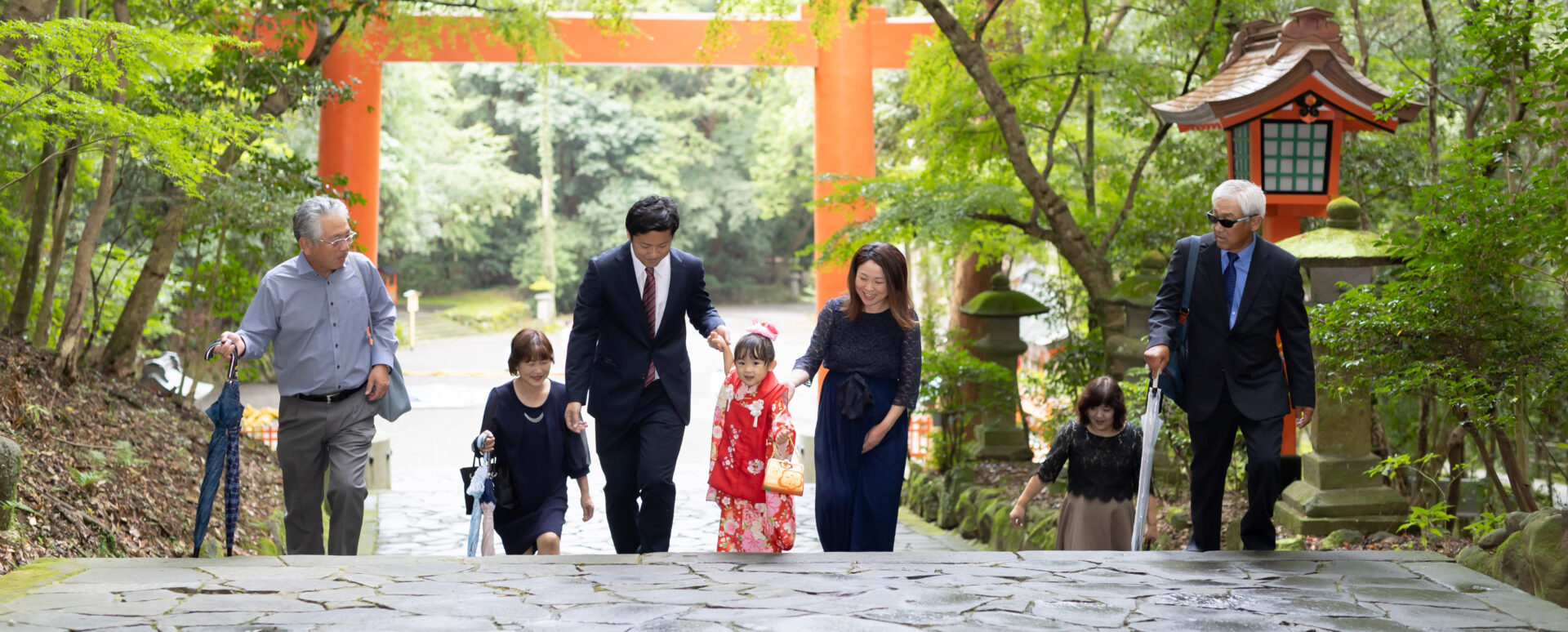 大分県宇佐市の宇佐神宮で七五三を迎えた3歳の女の子。赤い着物姿で、家族や祖父母と一緒に緑に囲まれた参道を歩く七五三のお参り写真。