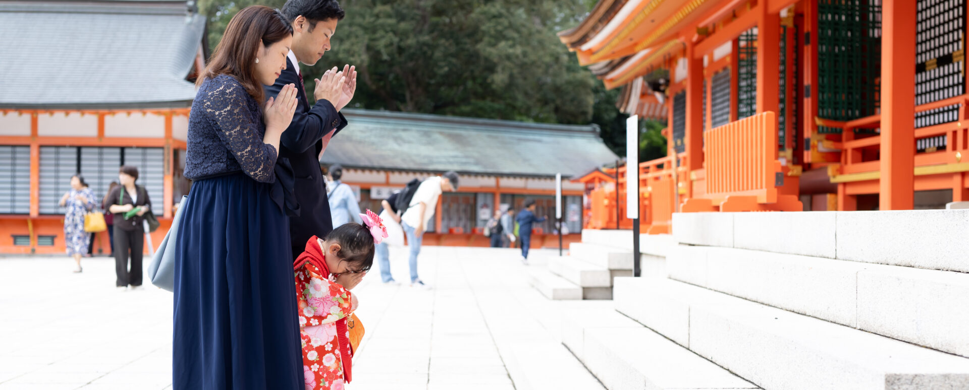 大分県宇佐市の宇佐神宮で七五三を迎えた3歳の女の子。赤い着物姿で、家族や祖父母と一緒に緑に囲まれた参道を歩く七五三のお参り写真。