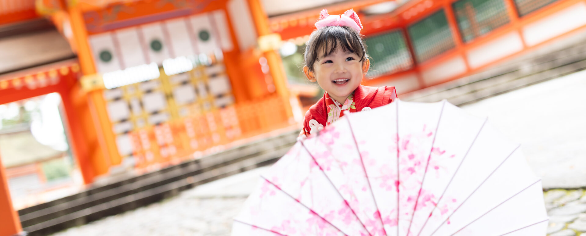大分県宇佐市の宇佐神宮で七五三のお参り。赤い着物姿の3歳の女の子が、和傘を持って楽しそうに笑う様子。