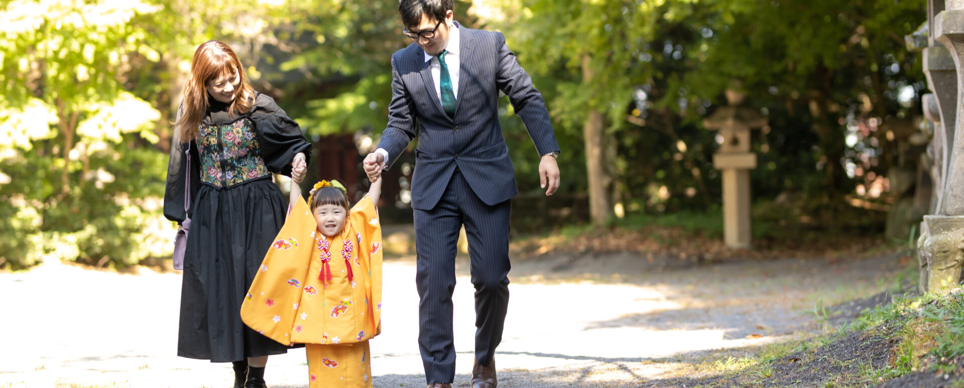 大分県中津市の薦神社で七五三のお参り。オレンジ色の着物姿の3歳の女の子が、パパとママと手をつないで楽しそうに歩く家族写真。