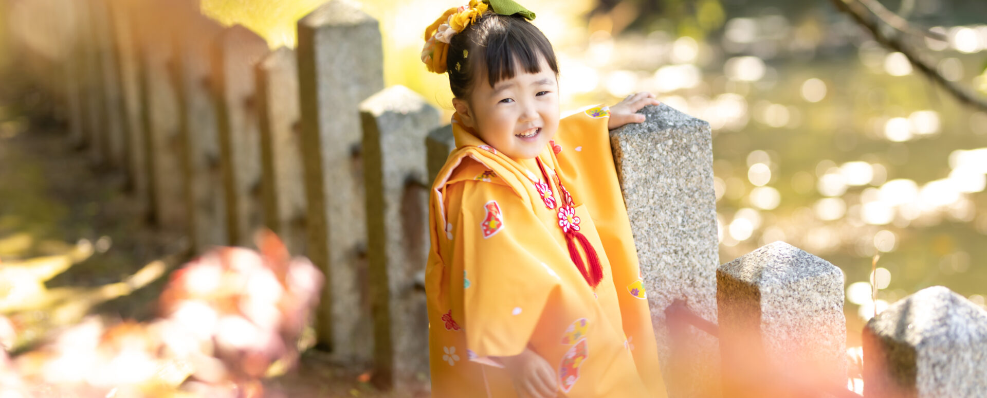 大分県中津市の薦神社で七五三のお参り。オレンジ色の着物姿の3歳の女の子が、境内の池のそばで楽しそうに笑う様子。