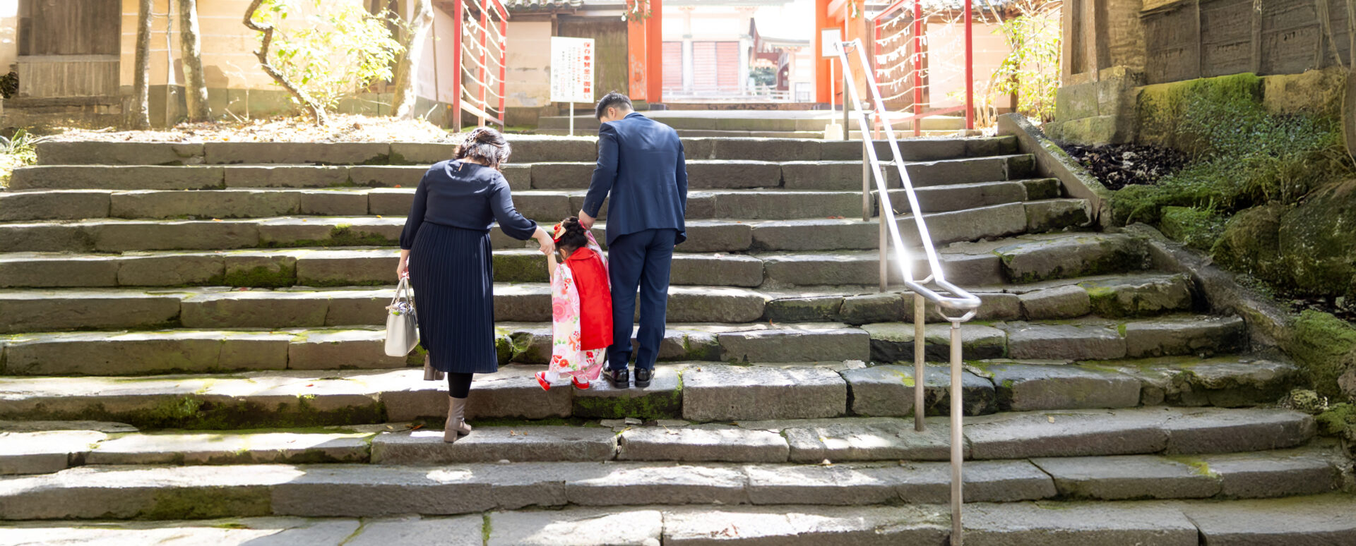 大分県大分市・春日神社での七五三。着物姿の女の子がパパとママに手をつないでもらい、歴史ある石段を一緒に上る様子。