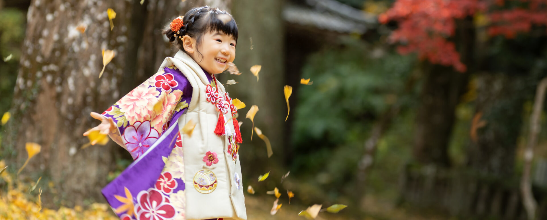 大分市の西寒多神社で七五三のお参りをする女の子。紅葉の中で落ち葉と遊びながら、楽しそうな笑顔を見せる記念写真。