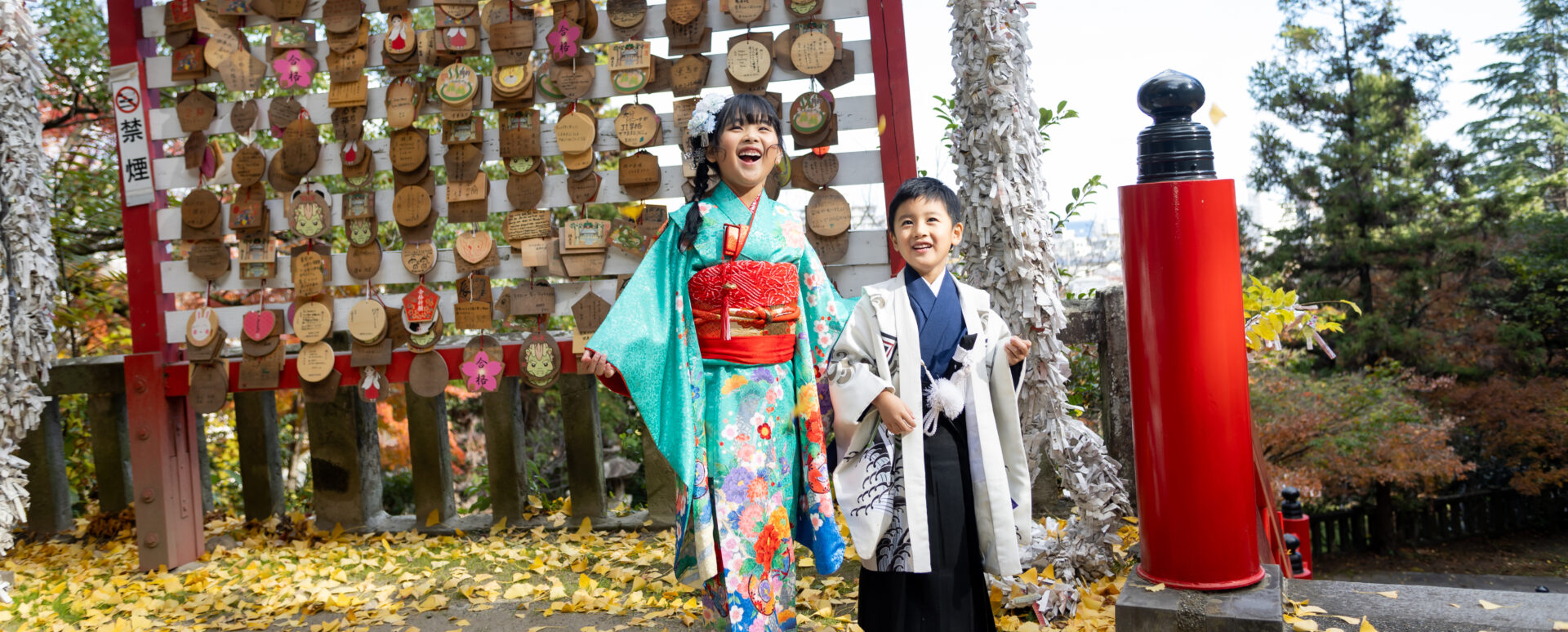 別府市の朝見神社で七五三を迎えた姉弟。色鮮やかな着物姿で、たくさんの絵馬と紅葉を背景に楽しそうに笑う七五三写真。