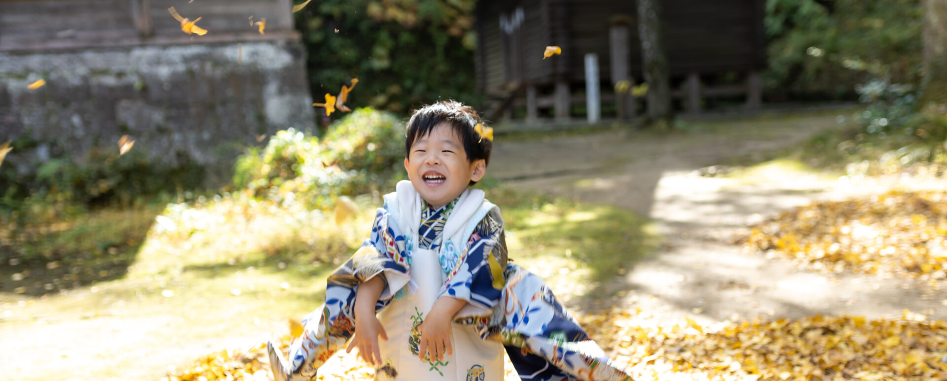 大分市の西寒多神社で七五三のお参りをする男の子。着物姿で落ち葉と遊びながら、元気いっぱいの笑顔を見せる記念写真。
