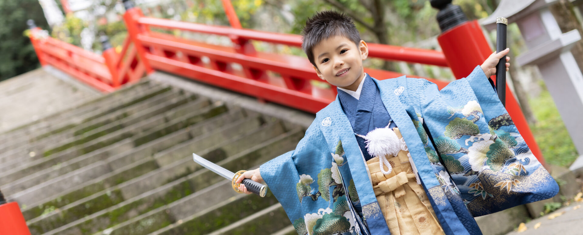 別府市の朝見神社で七五三を迎えた5歳の男の子。青い羽織袴姿で刀を手に持ち、赤い橋を背景に笑顔を見せる七五三写真。
