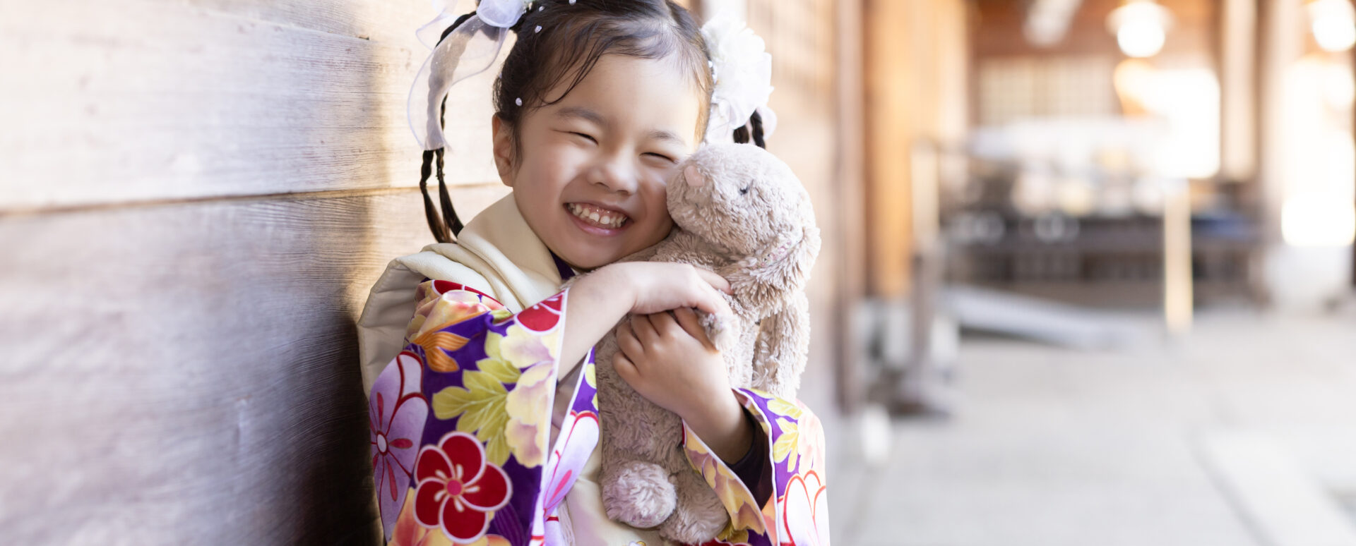 大分市の大分縣護國神社で七五三のお参りをする女の子。着物姿でお気に入りのぬいぐるみを抱きしめ、笑顔を見せる自然な記念写真。