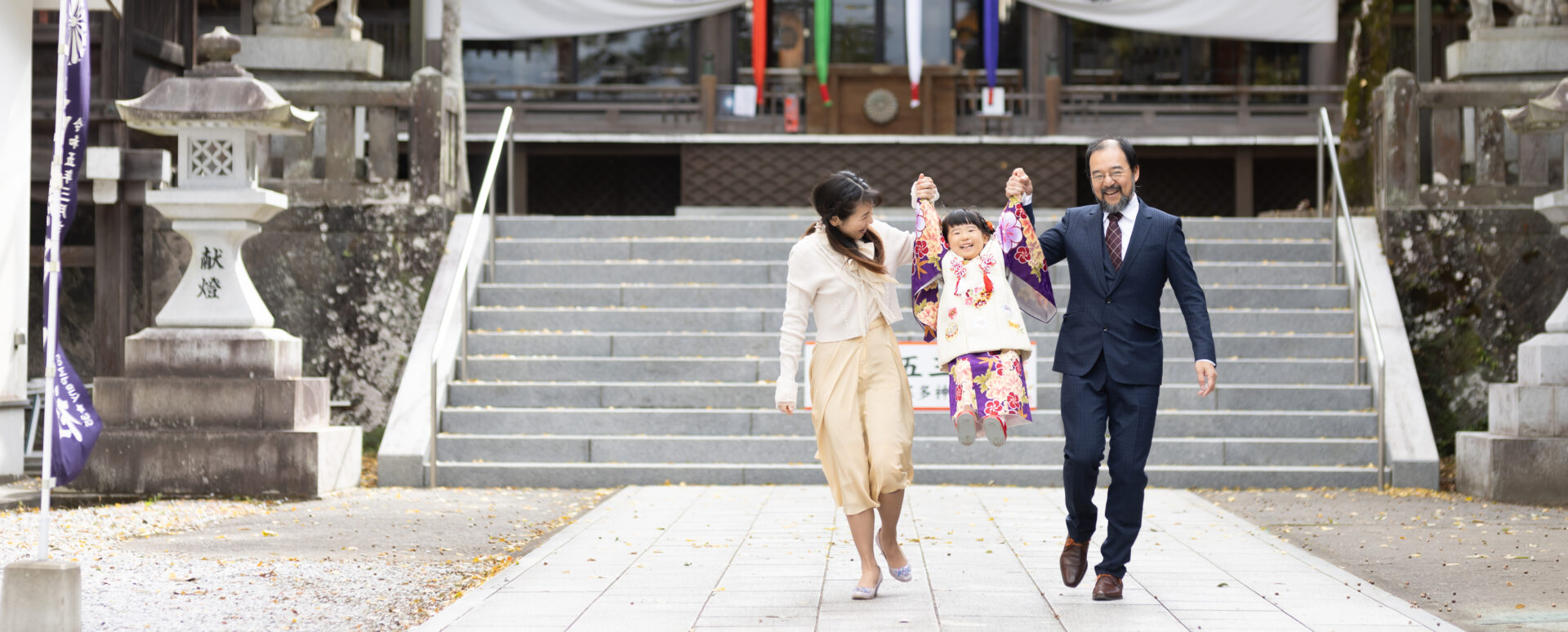 大分市の西寒多神社で七五三のお参りをする女の子。パパとママに両手をつないでもらい、笑顔で楽しむ自然な家族写真。