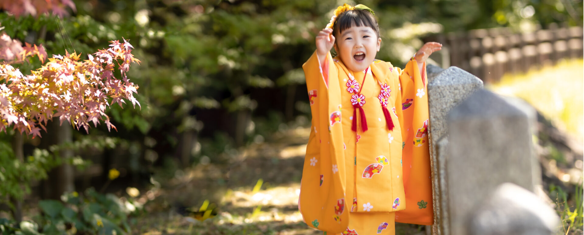 大分県中津市の薦神社で七五三のお参り。オレンジ色の着物姿の3歳の女の子が、紅葉の中で元気いっぱいに笑う様子。