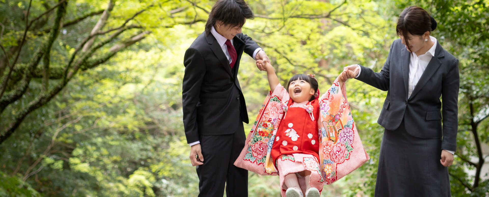 新緑に囲まれた神社で、パパとママに手をつないでもらい楽しそうに笑う3歳七五三のお参り同行撮影