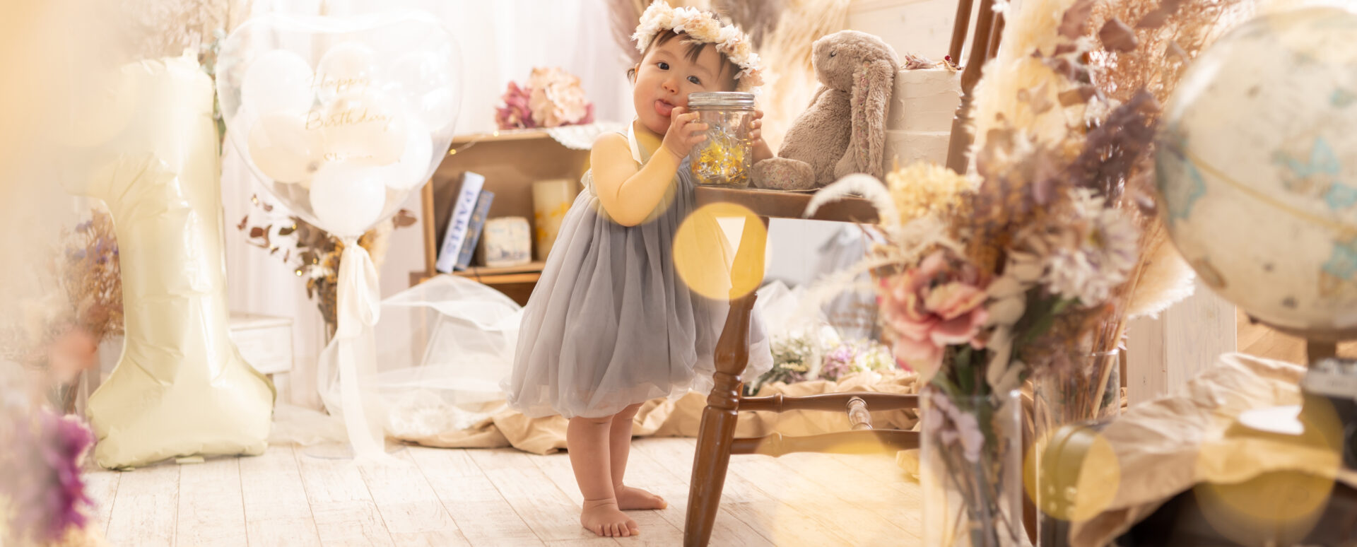 Photostudio PASSIONドライフラワーやパンパスグラス、1歳バルーンで飾られた明るい写真スタジオで、グレーのチュールドレスと花冠を着た1歳の女の子がつかまり立ちをして微笑むお誕生日記念写真