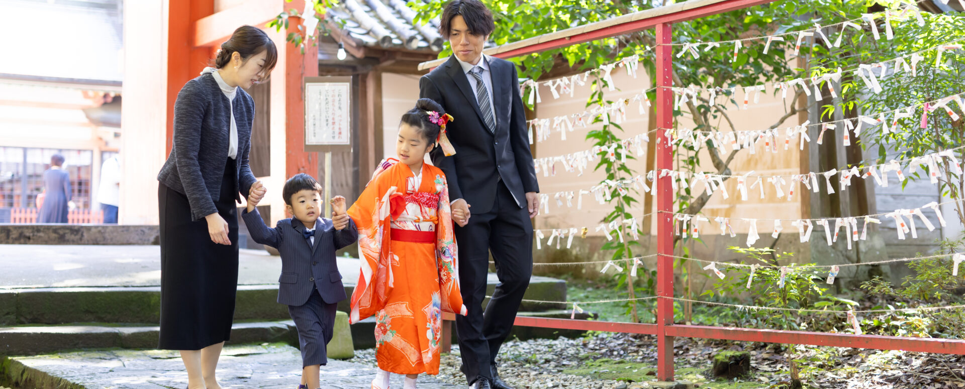 代替テキスト： 大分に柞原八幡宮で七五三のお参りで、オレンジ色の着物姿の女の子と弟がパパとママと手をつないで歩く家族写真