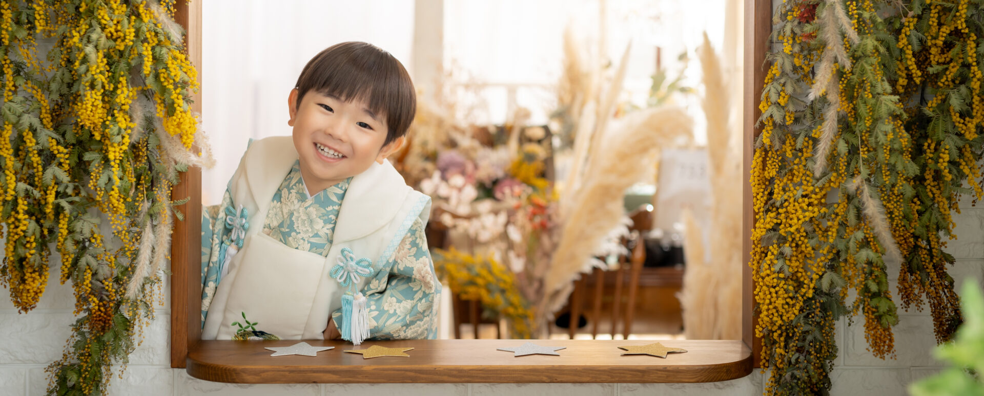 大分のPhotostudio PASSIONで、水色の着物と白い被布姿でミモザに囲まれ笑顔を見せる3歳七五三のスタジオ撮影
