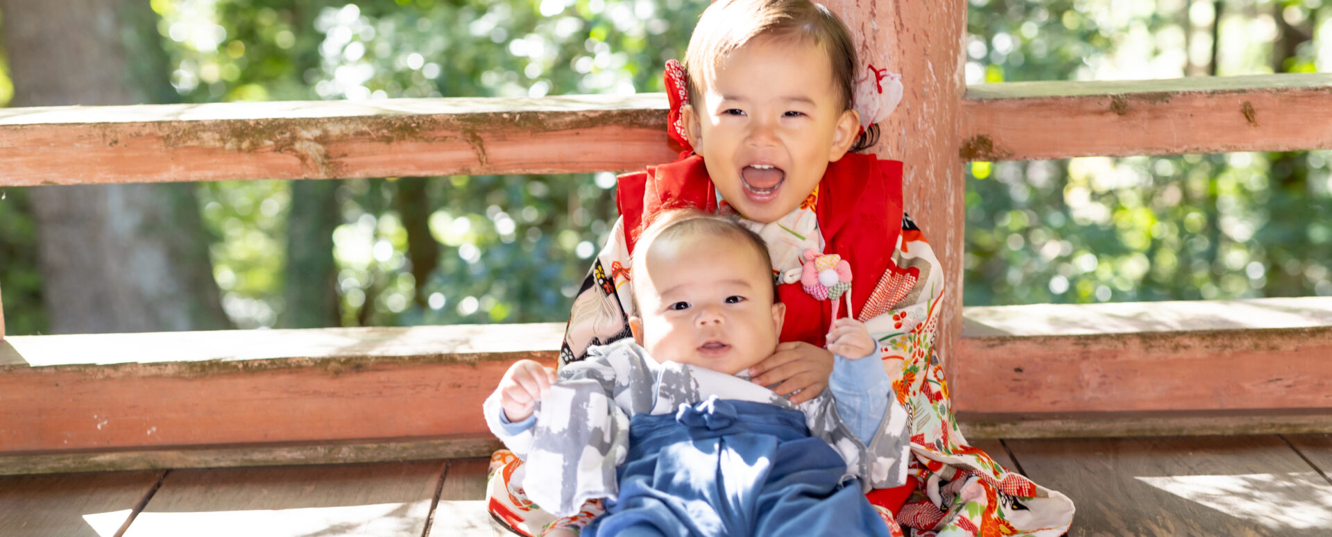 緑豊かな柞原八幡宮で、七五三の着物姿の女の子が和装の赤ちゃんを優しく抱く姉弟写真