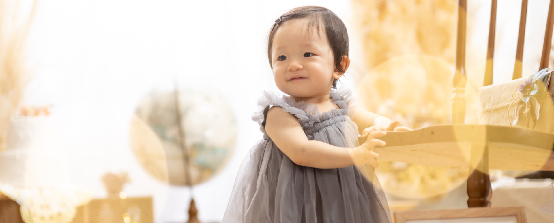 大分のPhotostudio PASSIONで、グレーのドレス姿でつかまり立ちをする1歳記念のスタジオ撮影